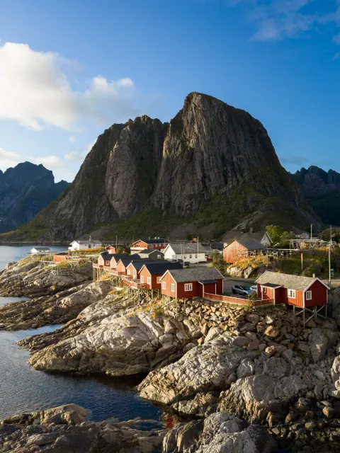 red-painted fishing cabins on a rocky shore below a steep granite peak, beside still water